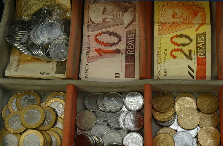 Uma gaveta de caixa registradora aberta revela cédulas e moedas brasileiras organizadas por valor, com notas de dez e vinte reais e moedas de diferentes denominações claramente visíveis.