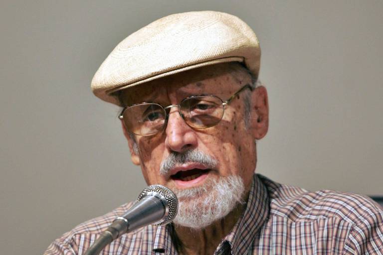 Homem idoso, de cavanhaque grisalho, utiliza boina branca e óculos, sentado atrás de uma mesa. Fotografado com expressão séria enquanto fala a um microfone posicionado à sua frente. 