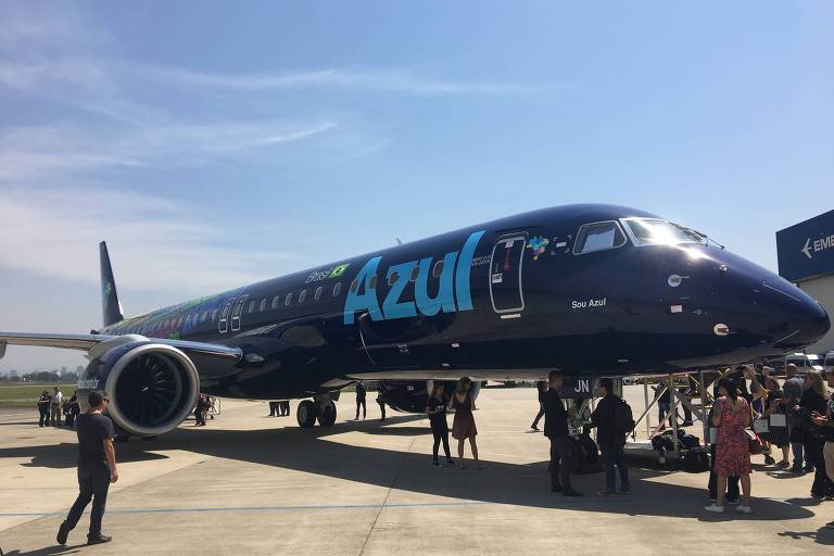 Um avião azul escuro, com o nome da companhia aérea em azul claro  é visto parado em um aeroporto. Várias pessoas estão ao redor