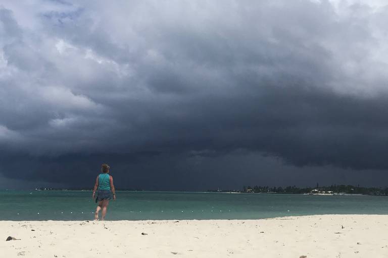 Uma mulher de shorts e camiseta está em pé na beira do mar, em uma praia de areias muito brancas. No mar, no horizonte, o céu está escuro, com nuvens carregadas, com a aproximação da tempestade Humberto. 