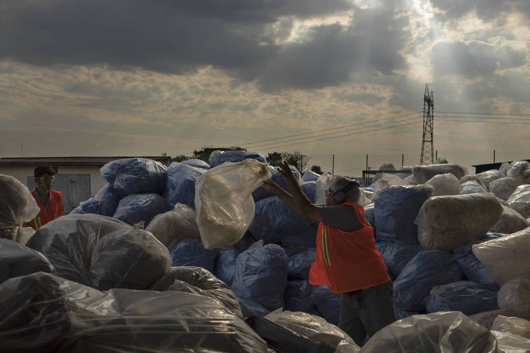 Homem vestido de laranja aparece em meio a vários sacos plásticos, em um depósito a céu aberto. 