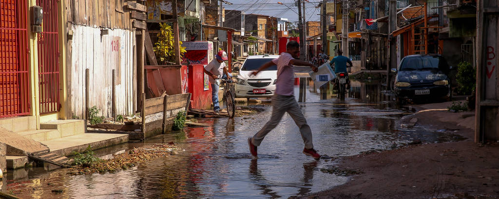 Pessoa atravessa rua repleta de água e lama, ladeada por casas de alvenaria e madeira