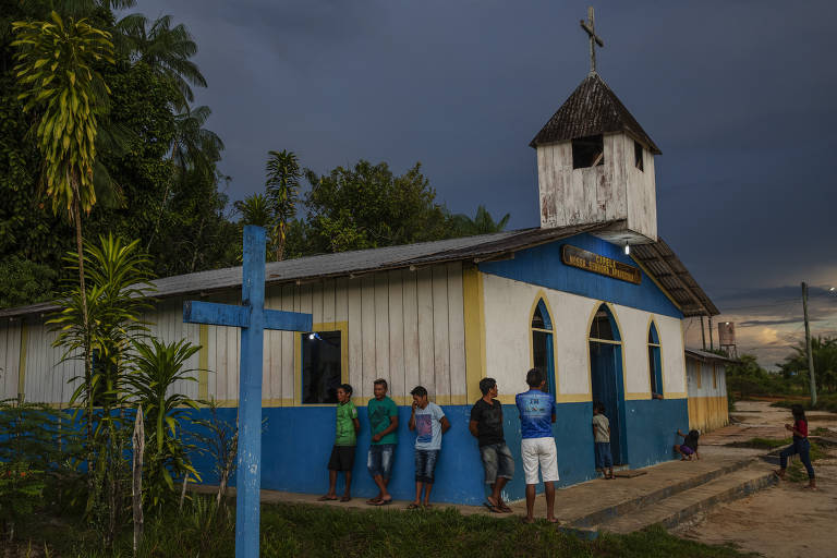 homens de chinelo camisa e bermuda do lado de fora de uma igreja de madeira nas cores branca e azul, com uma cruz de madeira azul fincada na terra à frente deles