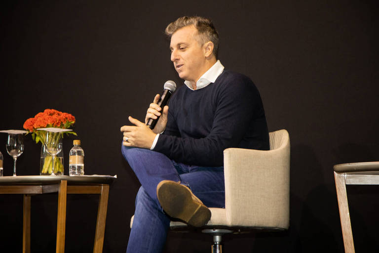 Luciano está sentado em uma cadeira, de pernas cruzas e com um microfone na mão; à sua esquerda há uma mesinha com flores e água