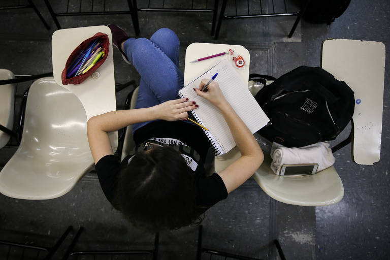 Sentada em carteira, aluna escreve em caderno; ao lado dela há uma mochila