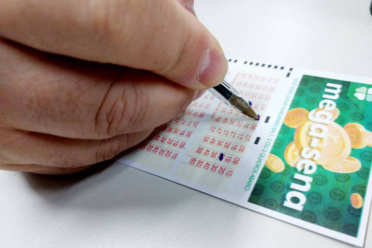 Uma mão marcando números em um volante da Mega-Sena com uma caneta sobre uma mesa