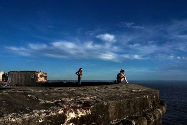 foto mostra grande pedra onde há uma mulher sentada. Atrás dela está uma outra mulher em pé. Há pessoas sentadas em cadeiras de praia, desfocadas. Ao fundo está o mar. O céu é azul e com poucas nuvens brancas