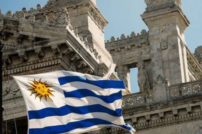 foto mostra bandeira do uruguai, que tem um sol com rosto em um quadrado de fundo branco do lado esquerod superior. O resto da bandeira é de faixas brancas e azuis na horizontal. Ao fundo, um  prédio de mármore