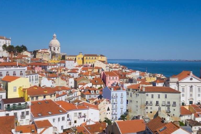 foto mostra diversas casas. A maioria tem paredes em cores claras, algumas azuis, outras amarelas e rosas. Todas possuem telhados laranjas. Ao fundo, do lado direito, está o mar.