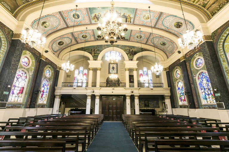 Interior de igreja, com tapete azul, bancos de madeira, lustres e paredes pintadas com afrescos 