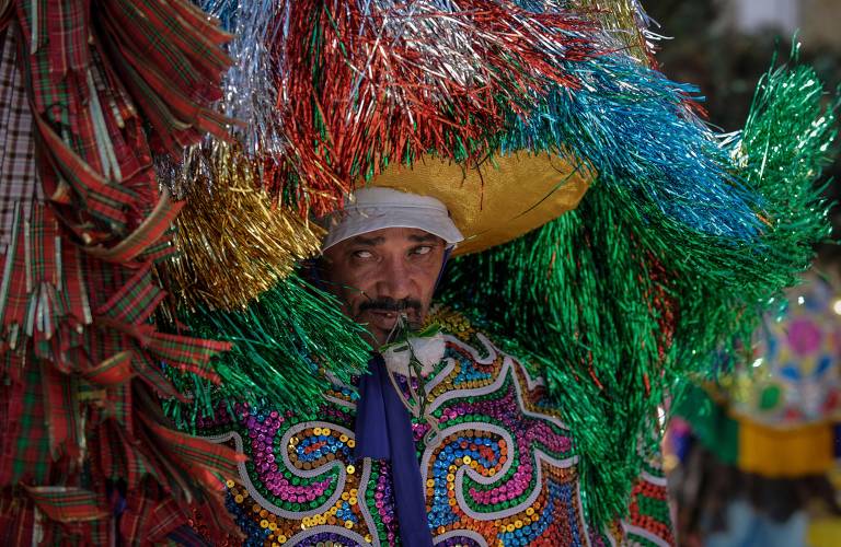 Homem usa chapeu e vestes coloridas de maracatu, com milhares de fitinhas coloridas 