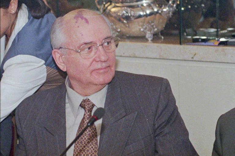 Gorbatchov aparece olhando para o lado, de óculos, frente a um microfone de mesa.
