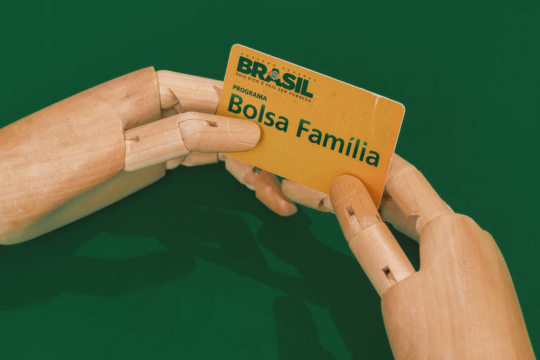 Foto mostra um fundo verde com duas mãos articuladas segurando um cartão amarelo do Bolsa Família.