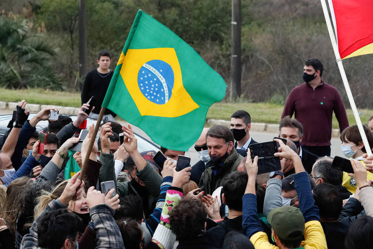 Bolsonaro cercado por por apoiadores, que apontam celulares e estendem as mãos. Um  deles estente uma bandeira do Brasil. O presidente usa máscar preta, mas o nariz e parte da boca estão descobertos