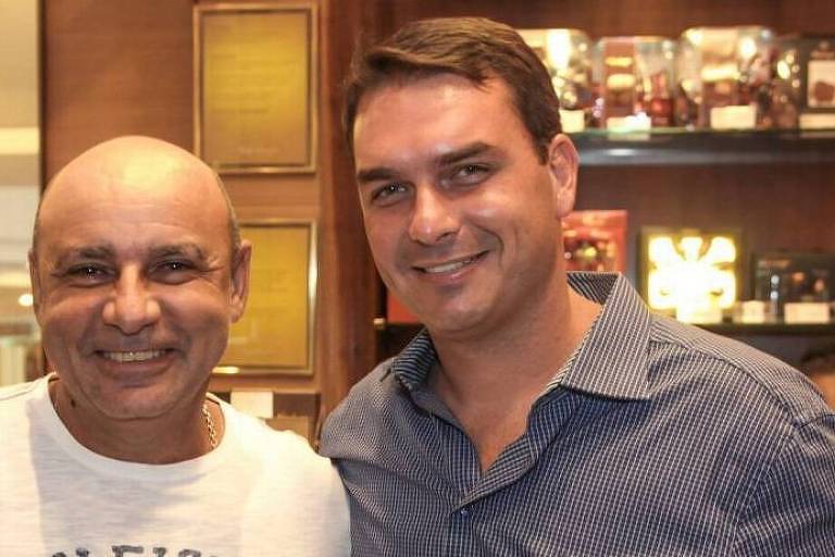 Fabrício Queiroz está à esquerda da foto. Ele é careca e veste camiseta branca. Está sorrindo. Flávio, do seu lado, é branco, tem cabelo castanho com um pequeno topete. Usa camisa em tom cinza e também sorri. Ao fundo, prateleiras com produtos