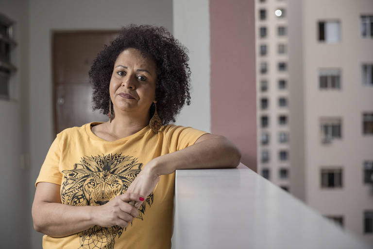 A assistente social Luciana Reis Oliviera, 48, em seu apartamento no centro de São Paulo; Luciana tem um meio sorriso na foto, em que aparece usando uma camiseta amarela, apoiada no parapeito do corredor do prédio, que é aberto