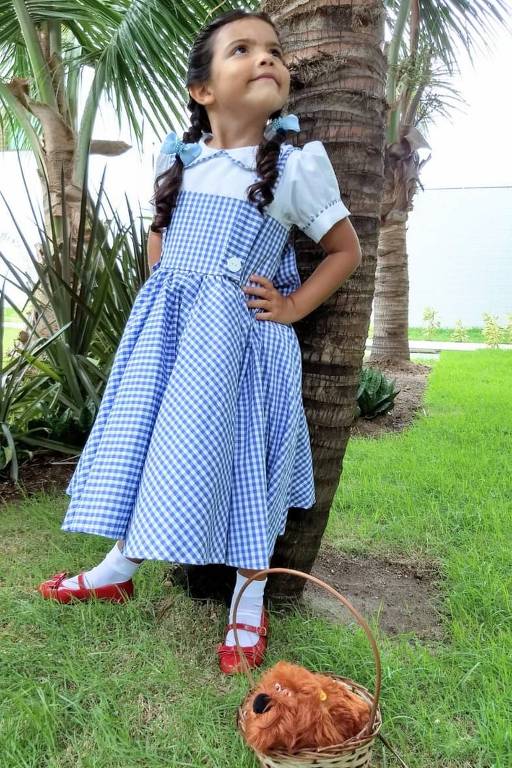 Criança branca usa vestido xadrez azul e branco e os sapatos vermelhos do filme. Ela está em pé na frente de uma árvore  