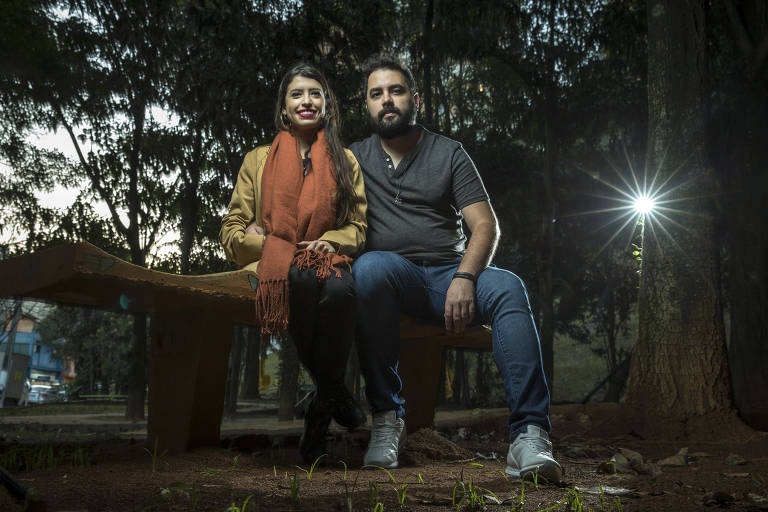 Homem e mulher sentados em banco de madeira, em praça, sorrindo 