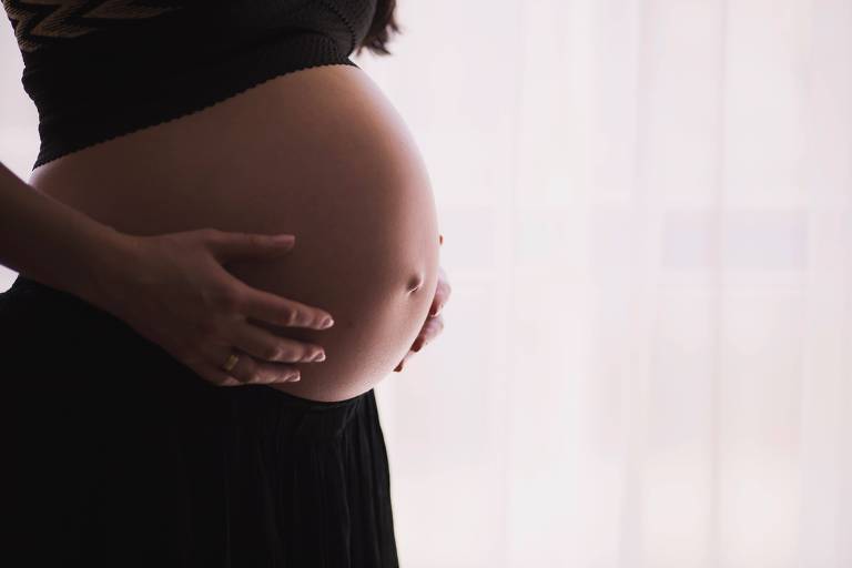 Mulher grávida segura a barriga, que está de fora da roupa. Imagem colorida