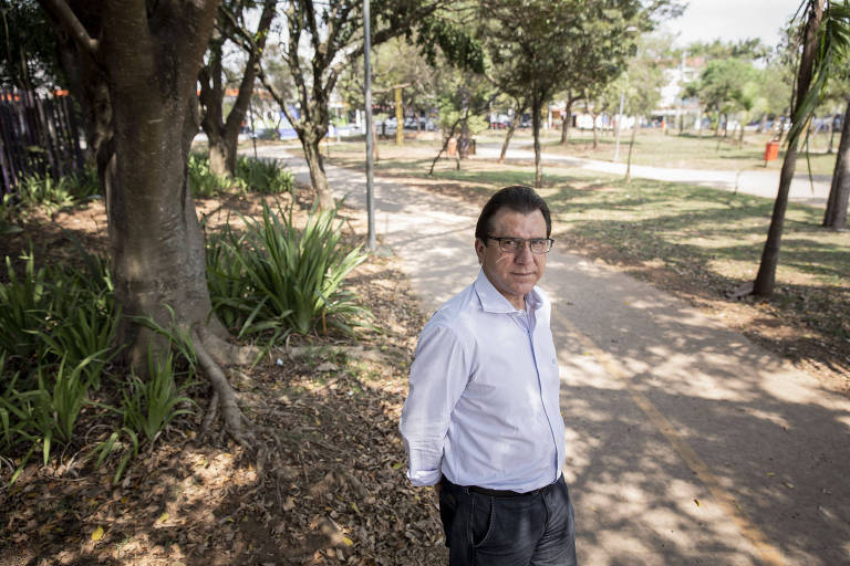 Um homem de camisa branca e óculos no meio de uma praça, com várias árvores e um caminho ao fundo