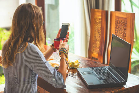 Mulher usando celular e laptop dentro de casa e bebendo café / chá.
Foto: astrosystem / Adobe stock