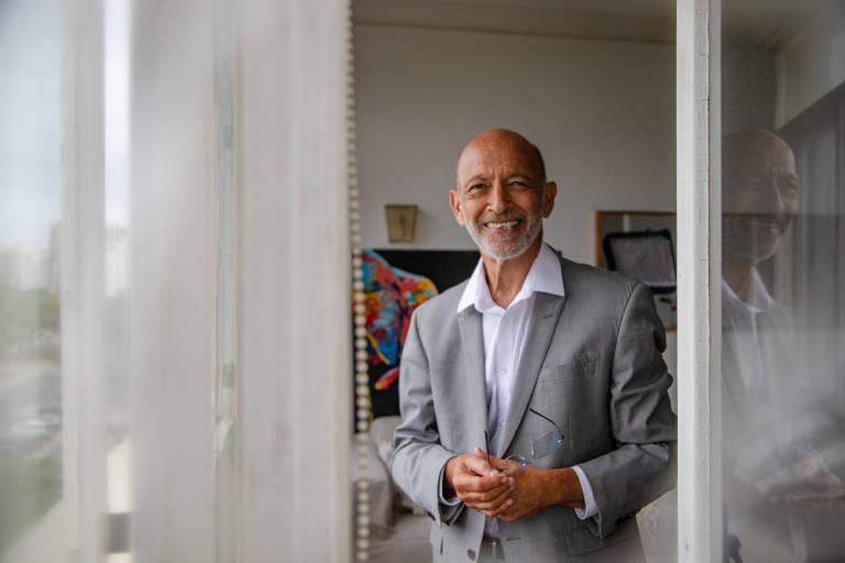 homem branco careca de terno cinza e camisa social posa para foto em uma sala com muitos quadros pendurados na parede 