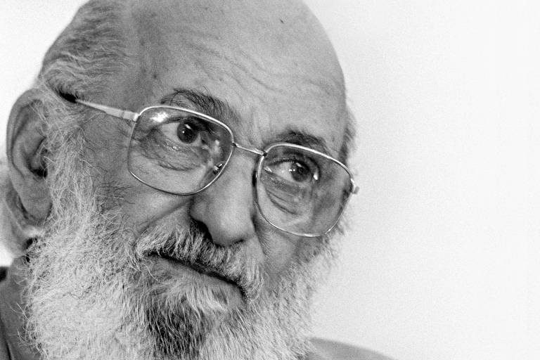 paulo freire tem uma longa barba branca e usa óculos, ele gesticula e olha para o lado 