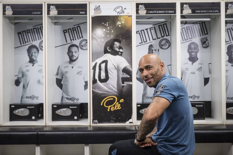 Edinho, filho de Pelé, de camiseta azul aparece diante da galeria de fotos ao lado do armário de Pelé, no vestiário do Santos no Estádio da Vila Belmiro