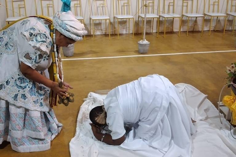 Uma mãe de santo está em frente a uma mulher ajoelhada aos seus pés, com roupas de candomblé