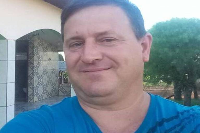 Em pose para selfie, Ênio Pasqualin, dirigente do MST no Paranáhomem de 48 anos, cabelos lisos, castanhos claros e camiseta azul. Ao fundo, aparece a fachada de uma casa e uma árvore.