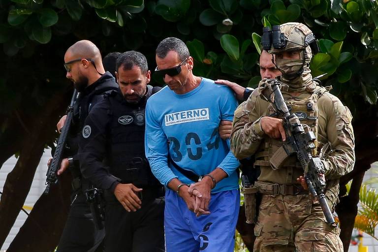 Marcola está no centro da foto, de roupa azul, óculos escuros e algemas nas mãos; ele está sendo escoltado por policiais. O que está a sua esquerda (à direita na foto) carrega uma arma nas mãos