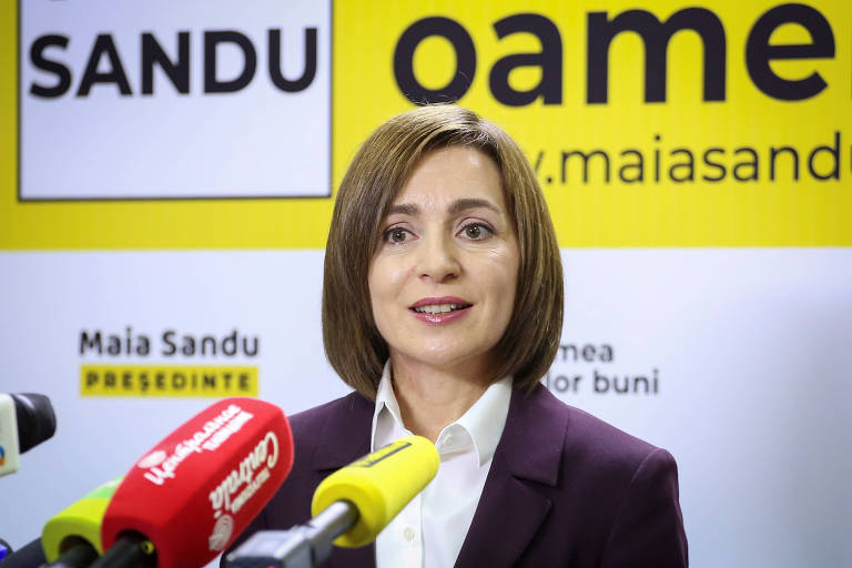 Mulher de cabelo curto, Maia Sandu, a presidente eleita da Moldova, com microfones à sua frente e um cartaz de propaganda política atrás