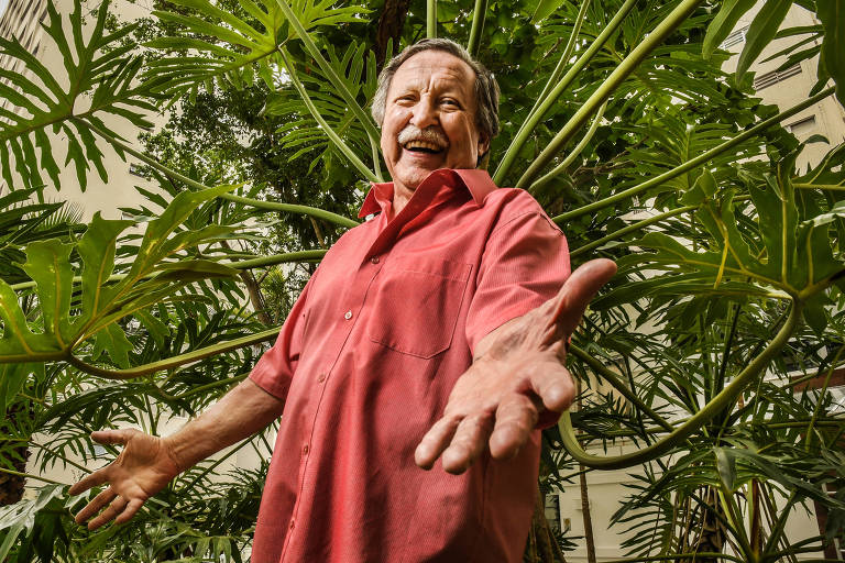 O escritor está de braços abertos sorrindo para  acâmera. Atrás dele, uma folhagem muito verde