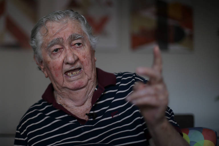 Homem branco de cabelo grisalho olha para a frente e gesticula com a mão esquerda enquanto conversa