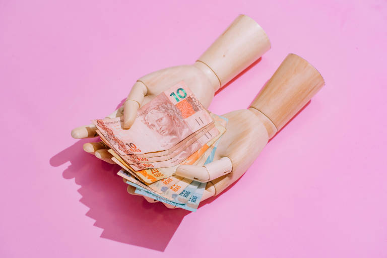 Prótese de mãos segura notas de dinheiro; fundo da foto é rosa