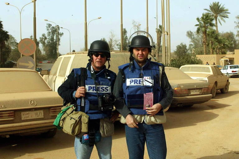 dois jornalistas em estacionamento coberto por terra; eles vestem coletes azuis à prova de bala e capacetes 