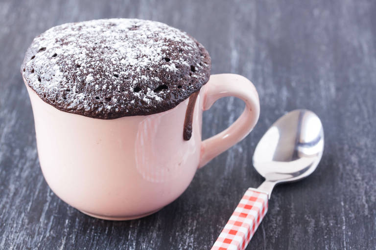 Bolo de chocolate em uma caneca cor de rosa, ao lado de uma colher. Estão sob uma mesa de madeira escura