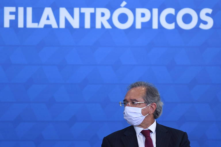 paulo guedes veste terno e está sentado, usando máscara, a frente de um painel azul com um letreiro branco em que se lê "filantrópicos" 