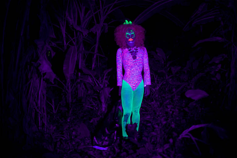 Foto escura, com luz negra, mostra pessoa com roupas e maquiagem neon, verdes e rosas, no centro