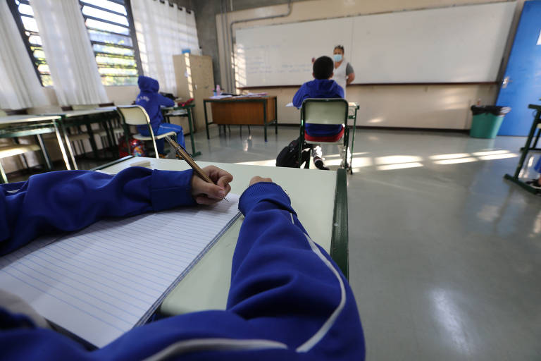 Dentro de uma sala de aula, imagem em close mostra os braços de uma criança sentada em uma carteira escolar apoiando os braços sobre a mesa e escrevendo em um papel. 