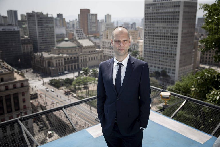 Bruno Covas é um homem branco e careca. Ele usa um terno, calça e gravata pretos, e uma camisa branca. Ao fundo, há a paisagem urbana da cidade de São Paulo.