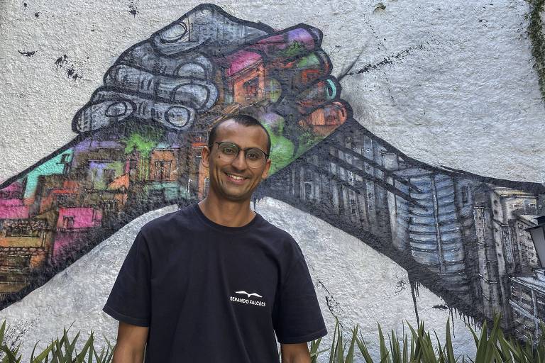 homem de camiseta preta e óculos posa em frente a muro grafitado colorido