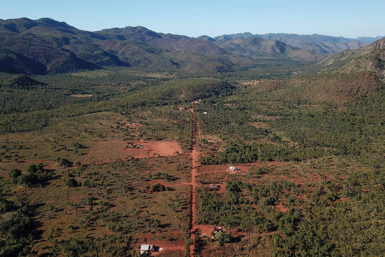 Paisagem rural de vegetação esparsa cortada por estrada de terra; o dia está ensolarado e há poucas casas vistas à distância