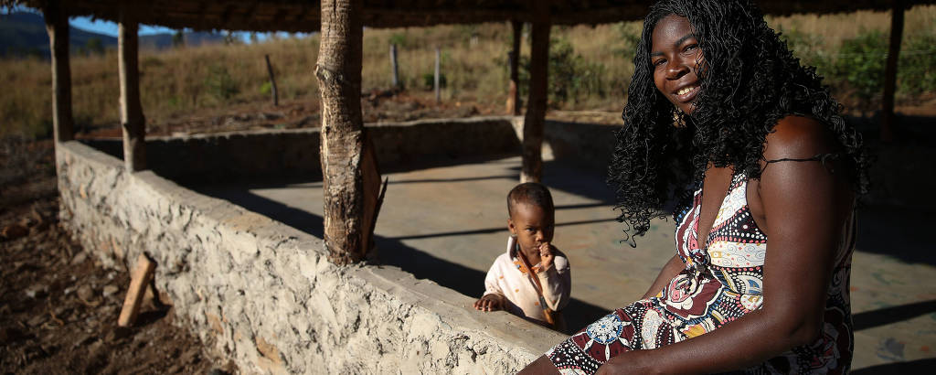 Em uma mureta da qual saem pilares de madeiras que sustentam o teto de palha, há uma mulher negra jovem, de cabelos longos e vestido estampado, sentada, olhando a câmera; um pouco atrás, há um menino, também negro, que aparenta três anos; é um dia de sol 