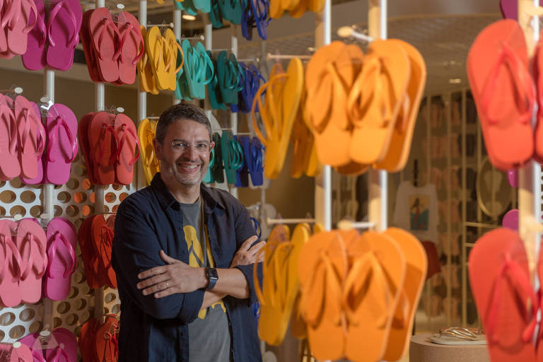 Homem branco de braços cruzados, sorrindo, ao lado de havaianas 