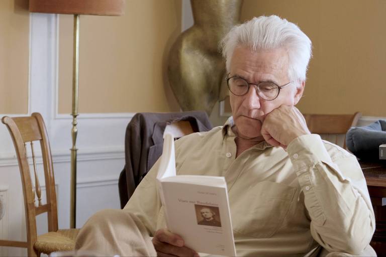 homem branco de cabelos brancos e óculos sentado lendo