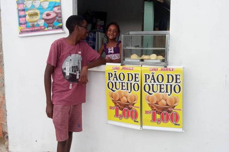 duas crianças posam em frente a uma janela de casa com cartaz sobre venda de pães de queijo