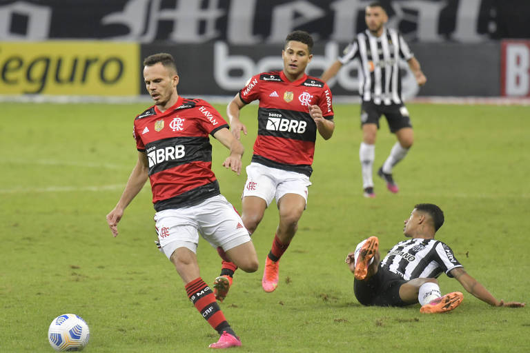 Dois homens de vermelho e preto correndo com bola em gramado, com dois homens de preto e branco atrás 