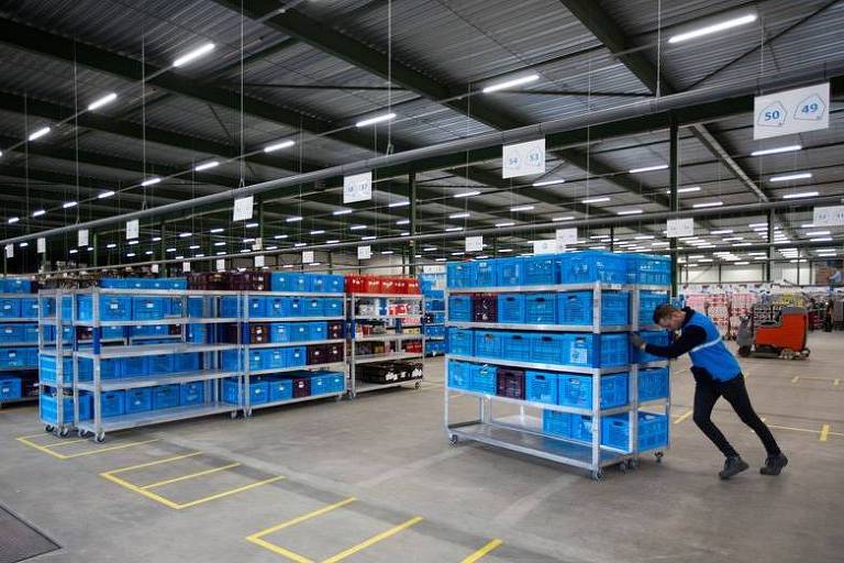 Imagem mostra funcionário, vestindo uniforme azul e calça preta, empurrando carrinho cheio de caixas azuis, dentro de um galpão. Nele, há vários outros carrinhos com as mesmas caixas.