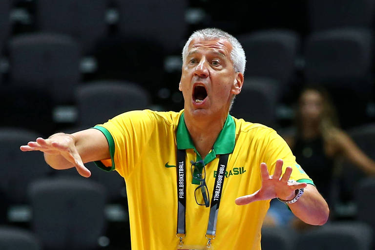 Um treinador de voleibol de cabelos grisalhos está gesticulando com as mãos enquanto dá instruções. Ele veste uma camisa amarela com detalhes verdes e usa um crachá pendurado no pescoço. Ao fundo, há cadeiras vazias e uma pessoa desfocada.
